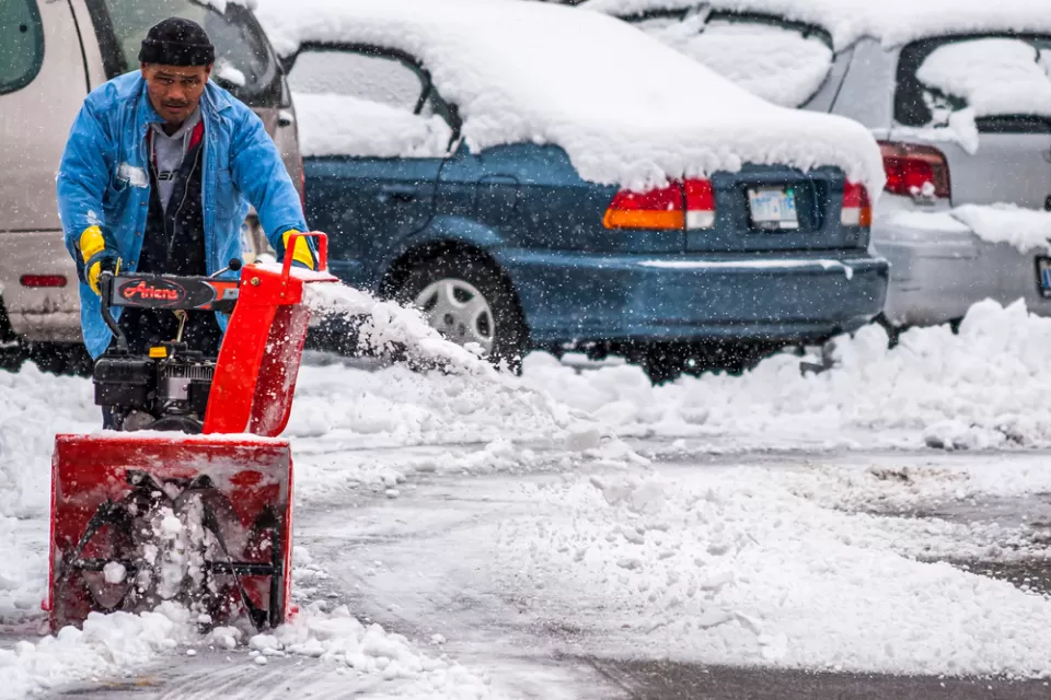A man pushes a snow blower to clear the ground of an outdoor parking lot filled with cars.